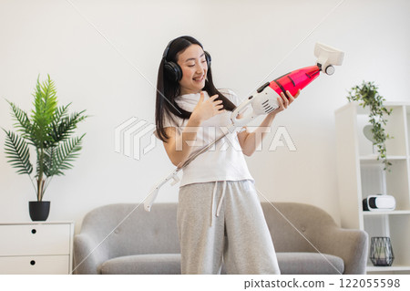 Asian young woman wearing headphones joyfully cleaning living room with vacuum, embracing fun in household chores. 122055598