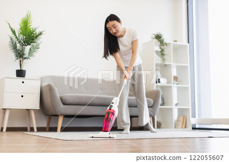 Asian woman in casual clothing vacuuming living room floor. Scene depicts cleanliness, home care, and modern lifestyle. Bright room with minimalist decor adds to contemporary feel. Asian woman in casual clothing vacuuming living room floor. Scene depicts cleanliness, home care, and modern lifestyle. Bright room with minimalist decor adds to contemporary feel. 122055607
