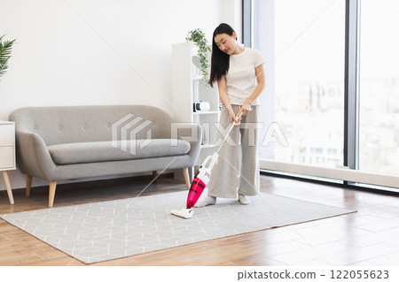 Young Asian woman cleaning living room with vacuum cleaner. Modern bright interior with sofa and plants. Concept of home cleanliness and domestic chores. 122055623