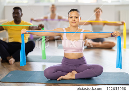 Asian woman sitting performs exercises with wide elastic band during Pilates multinational workout. Asian woman sitting performs exercises with wide elastic band during Pilates multinational workout. 122055968