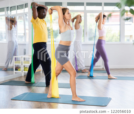 Girl conducts Pilates training for multinational students, standing exercises with wide elastic band Girl conducts Pilates training for multinational students, standing exercises with wide elastic band 122056010