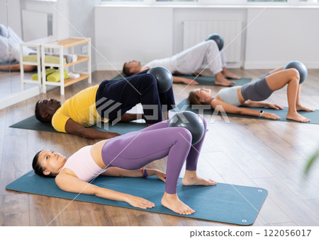 Multinational group of people lie on mats, hold Pilates ball between their knees and perform abdominal exercises during group exercise in fitness studio Multinational group of people lie on mats, hold Pilates ball between their knees and perform abdominal exercises during group exercise in fitness studio 122056017