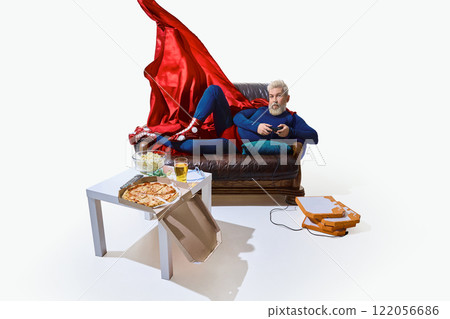Man in superhero costume with red cape, lounging on couch with beer and snack, playing video game with game controller against white studio background 122056686