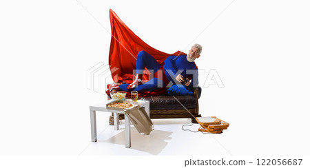 Man in superhero costume with red cape, lounging on couch with beer and snack, playing video game with game controller against white studio background 122056687
