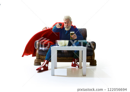 Evening chill at home, Man in superhero costume sitting on couch with remote TV controller, drinking beer, table filled with popcorn against white studio background Evening chill at home, Man in superhero costume sitting on couch with remote TV controller, drinking beer, table filled with popcorn against white studio background 122056694