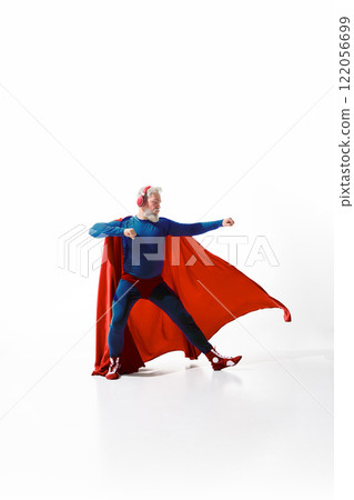 Dynamic image of grey-haired man superhero costume with red cape, listening to music in headphones and standing with fist up against white studio background 122056699