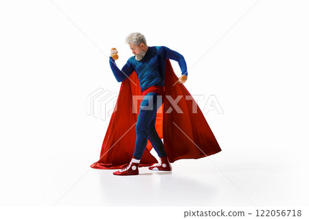 Grey-haired athletic man in superhero costume with red cape standing in dynamic pose with dumbbells, against white studio background Grey-haired athletic man in superhero costume with red cape standing in dynamic pose with dumbbells, against white studio background 122056718