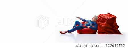 Man in superhero costume lounging on red beanbag with TV remote control against white studio background. 122056722