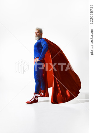 Gray-haired man in blue superhero outfit and red cape stands tall and determined, showing strength and resilience against white studio background. 122056735
