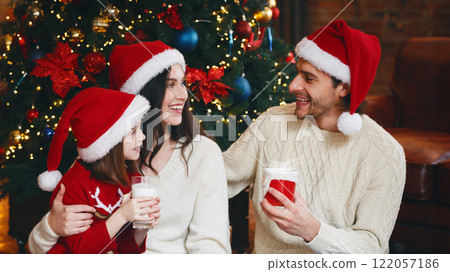 Sweet young happy family drinking warm milk on Christmas eve next to xmas tree, having conversation 122057186