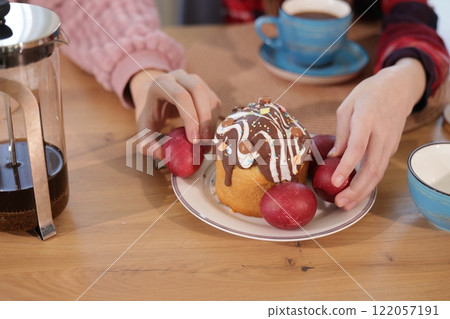 Hands arranging red dyed eggs around a decorated Easter cake topped with chocolate icing on a wooden table, with a French press and a blue cup nearby. 122057191
