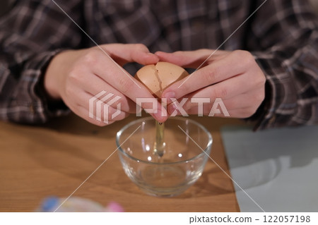 Close-up of hands cracking an egg over a glass bowl, preparing ingredients for baking on a wooden table. Close-up of hands cracking an egg over a glass bowl, preparing ingredients for baking on a wooden table. 122057198