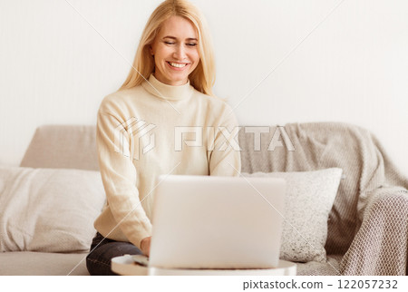 A woman sits on a couch, smiling and looking down at a laptop in her lap. A woman sits on a couch, smiling and looking down at a laptop in her lap. 122057232