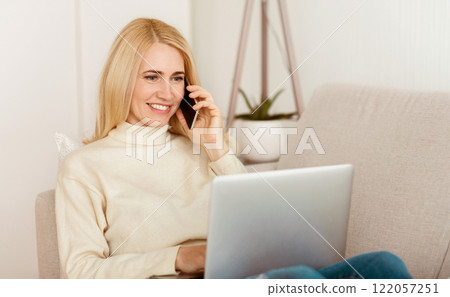A woman sits on a couch, smiling while talking on a cell phone and working on her laptop. A woman sits on a couch, smiling while talking on a cell phone and working on her laptop. 122057251