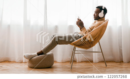 Weekend. Relaxed Black Man In Wireless Headset Using Mobile Phone Listening To Audiobook Sitting On Modern Chair Against Window Indoor 122057268