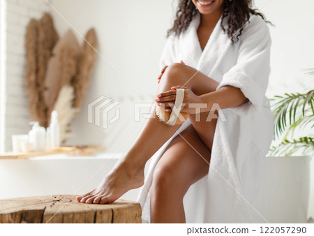 Unrecognizable Black Woman Dry Brushing Legs Making Anti-Cellulite Massage Sitting On Bathtub In Bathroom Indoor, Wearing Bathrobe. Body Care Routine. Cropped Shot, Selective Focus 122057290