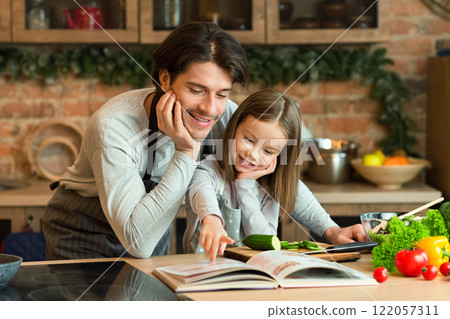 Caring young dad and little daughter searching recipe in culinary book, preparing healthy dinner together in kitchen, copy space 122057311