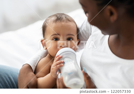 Nursery And Feeding Concept. Closeup of adorable small black baby drinking from bottle, young smiling woman holding kid on hands, taking care of her child, sitting on bed at home, selective focus Nursery And Feeding Concept. Closeup of adorable small black baby drinking from bottle, young smiling woman holding kid on hands, taking care of her child, sitting on bed at home, selective focus 122057330