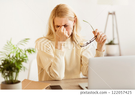 A woman sits at a desk, rubbing her eyes and holding her glasses. She looks tired and may be experiencing eye strain from working on her laptop. 122057425