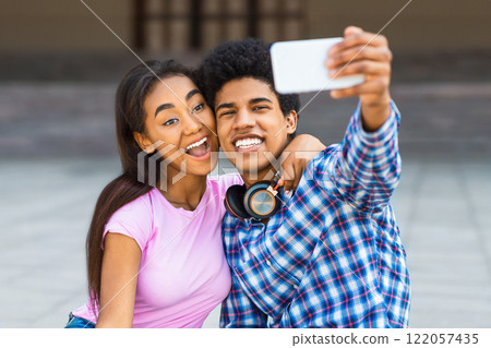 A teen black couple smiles for a selfie in the city. Guy is wearing a plaid shirt and headphones around his neck. The girl is wearing a pink shirt and jeans. A teen black couple smiles for a selfie in the city. Guy is wearing a plaid shirt and headphones around his neck. The girl is wearing a pink shirt and jeans. 122057435