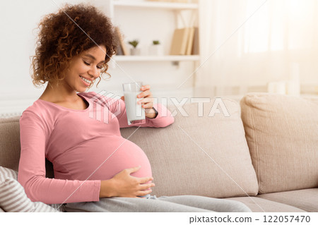 Healthy pregnancy. African expectant woman holding glass of fresh milk, sitting on sofa, empty space 122057470