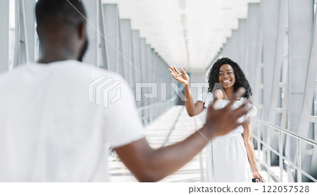 African American woman in a white dress is waving goodbye to a man as she walks away from him in an airport terminal. The man is wearing a white t-shirt and is looking towards the woman. 122057528