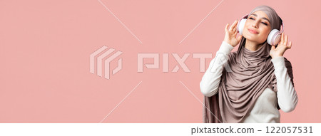 Joyful muslim girl in headscarf listening favorite music with wireless headphones, standing over pink background with copy space Joyful muslim girl in headscarf listening favorite music with wireless headphones, standing over pink background with copy space 122057531