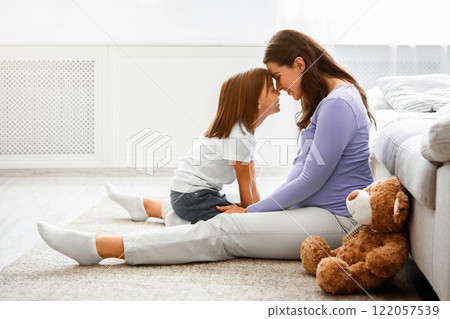 Family and love concept. Adorable little girl and expecting mother cuddling at home, sitting on floor, touching each other with foreheads, copy space 122057539