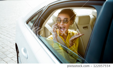 A woman with curly hair and sunglasses is sitting in the back seat of a white car. She is on a phone call and looks excited, with her mouth open in surprise. 122057554