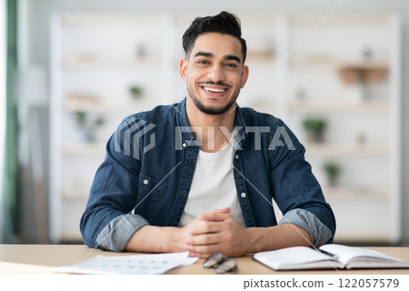 Cheerful arab guy in casual sitting at workdesk and smiling at camera, office interior, copy space. Happy young middle-eastern man sitting at table with notebook and papers, home interior Cheerful arab guy in casual sitting at workdesk and smiling at camera, office interior, copy space. Happy young middle-eastern man sitting at table with notebook and papers, home interior 122057579