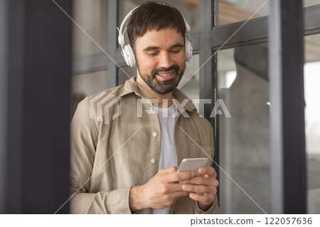 A man in a tan shirt and white t-shirt is standing in an office setting. He is wearing headphones and looking down at his smartphone. He appears to be listening to something on his phone. 122057636