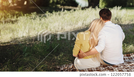 Couple sitting in summer park, having picnic on blanket. Lovers hugging and looking at lake 122057758