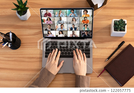 A person is participating in a video conference using a laptop computer. The screen shows multiple people in a grid format. The persons hands are on the keyboard of the laptop, top view A person is participating in a video conference using a laptop computer. The screen shows multiple people in a grid format. The persons hands are on the keyboard of the laptop, top view 122057775