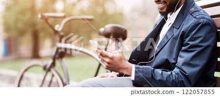 A businessman is seated on a park bench, using his smartphone, with a bicycle nearby. The environment is serene and professional, showcasing a balance between work and nature, cropped 122057855