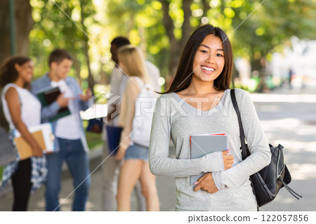 Happy asian girl student holding notepad and smiling at camera, spending time with friends in public park 122057856