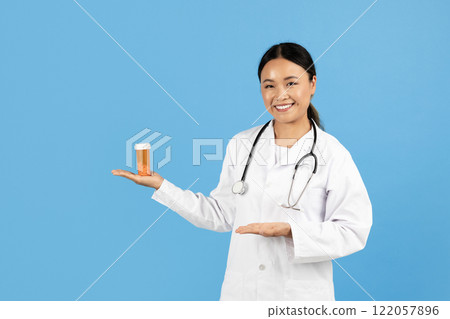 A smiling Asian lady healthcare professional in a white coat presents a prescription bottle while standing against a blue background. A smiling Asian lady healthcare professional in a white coat presents a prescription bottle while standing against a blue background. 122057896