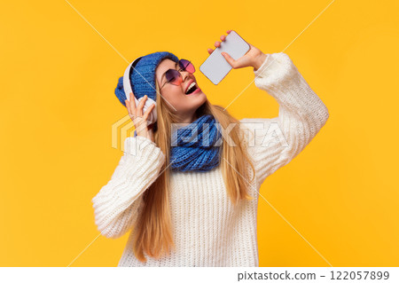Joyful young winter girl in wireless headphones listening to music and singing in phone over pink background 122057899