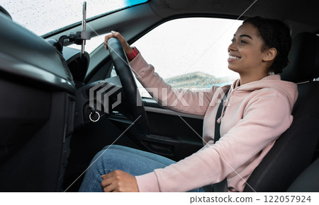 Great friendly taxi driver, auto blogger and car traveler. Smiling millennial african american woman holding steering wheel, changing speed, looking at road, driving car in rain, profile, empty space 122057924