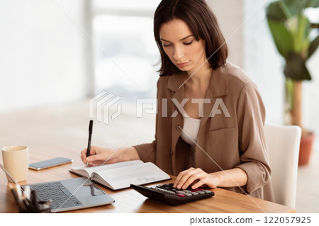 Budget Planning. Focused young woman using calculator, counting income and expenses and writing, free space 122057925