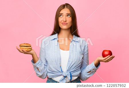 Puzzled Girl Choosing Between Burger And Apple Standing Over Yellow Studio Background. Healthy Vs Unhealthy Food. Puzzled Girl Choosing Between Burger And Apple Standing Over Yellow Studio Background. Healthy Vs Unhealthy Food. 122057932