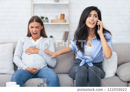 Preterm Labor. Girl Calling Doctor For Her Pregnant Friend Having Spasm Suffering From Pain Sitting On Sofa Indoor. Preterm Labor. Girl Calling Doctor For Her Pregnant Friend Having Spasm Suffering From Pain Sitting On Sofa Indoor. 122058022