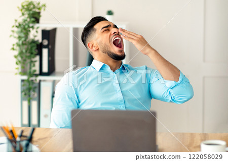 Sleepy young Arab guy yawning, tired of working with laptop at modern office. Millennial workaholic suffering from chronic fatigue, feeling sleepy or bored at workplace, copy space Sleepy young Arab guy yawning, tired of working with laptop at modern office. Millennial workaholic suffering from chronic fatigue, feeling sleepy or bored at workplace, copy space 122058029