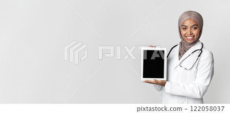 Online pharmacy concept. Black muslim female doctor holding digital tablet with black blank screen for mockup, posing over gray background, free space 122058037