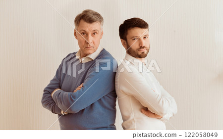 Relationship Problems. Serious Adult Son And Senior Father Standing Back-To-Back Crossing Hands Against White Wall Indoor 122058049