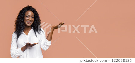 African American smiling young woman with curly black hair and brown skin, dressed in a white button-up shirt, standing in front of a peach-colored background. She is extending her right hand African American smiling young woman with curly black hair and brown skin, dressed in a white button-up shirt, standing in front of a peach-colored background. She is extending her right hand 122058125