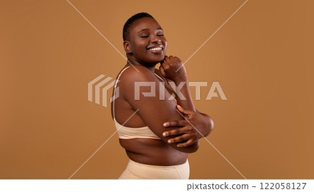 Self Acceptance, Love And Care. Portrair of half-turned smiling curvy African American woman with perfect skin and closed eyes embracing herself isolated on brown studio background, free copy space 122058127