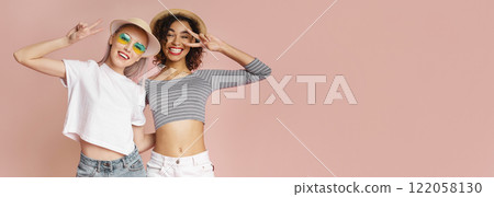 This photo shows two young women in hats smiling and making peace signs. They are standing in front of a pink wall, and both women are looking at the camera, copy space 122058130