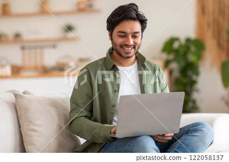 Arab man is sitting on a white couch in a modern home. He is smiling and looking at his laptop. He is wearing a green shirt, blue jeans, and a white t-shirt. There is a plant in the background. 122058137