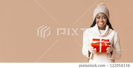 Happy african woman in white winter hat and gloves holding Christmas present, beige studio background, copy space 122058138