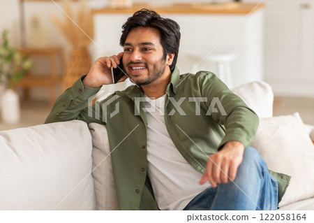 A young Arab man with a beard, wearing a green shirt and blue jeans, sits on a white couch and talks on his phone while smiling. A young Arab man with a beard, wearing a green shirt and blue jeans, sits on a white couch and talks on his phone while smiling. 122058164